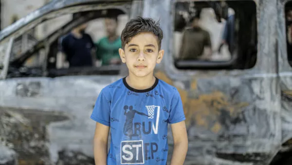 A young boy standing in front of a burnt car in Gaza. 