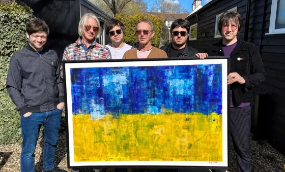 Andy Crofts and Paul Weller band hold up the artwork being auctioned off. The artwork resembles the Ukrainian flag with the top half painted blue and bottom half painted yellow.