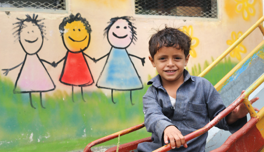 Boys sits on slide outside War Child child-friendly space in Yemen, in front of a wall painted with brightly coloured stick figures.