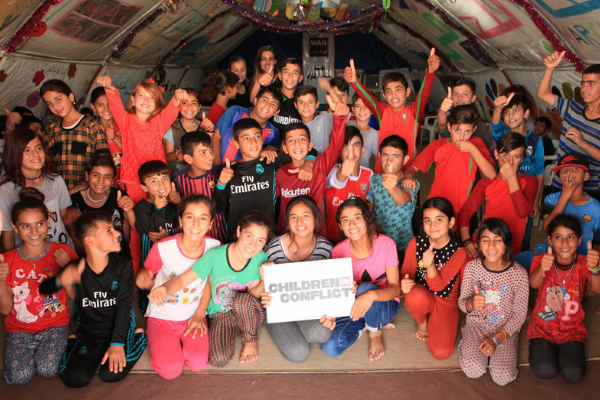 Children in a displacement camp hold up a sign with Children in Conflict's Logo on it.