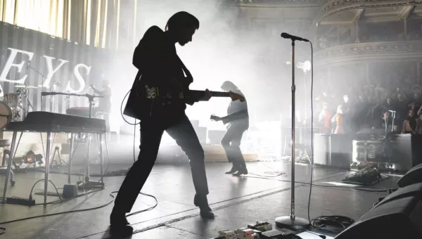 Arctic Monkeys live on stage. Image is black and white with lead singer as a silhouette in the foreground with bright white lights behind. 