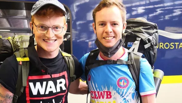 Mitch Jones wearing a War Child branded t-shirt and baseball cap smiles to the camera stood in front of a Eurostar train.