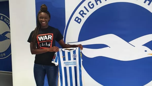 Rinsola Babjide stands in front of a wall covered in blue and white striped and the Brighton & Hove Football Club logo whilst holding her blue and white striped Brighton & Hove Football jersey. Rinsola wears a black War Child T-Shirt with War Child logo on the front.