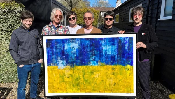 Andy Crofts and Paul Weller band hold up the artwork being auctioned off. The artwork resembles the Ukrainian flag with the top half painted blue and bottom half painted yellow.