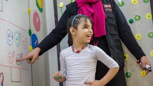 Girl smiles to her classmates as she completes a task on the whiteboard with the help of a War Child Education Facilitator in Jordan.