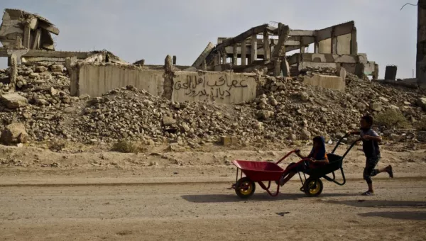 A child pushing a wheel barrow next to a destroyed building. 