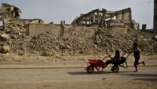 A child pushing a wheel barrow next to a destroyed building.