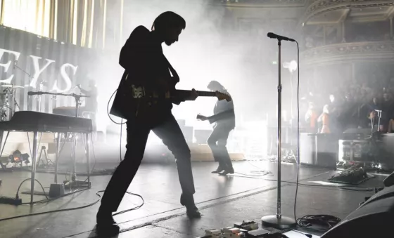 Arctic Monkeys live on stage. Image is black and white with lead singer as a silhouette in the foreground with bright white lights behind. 