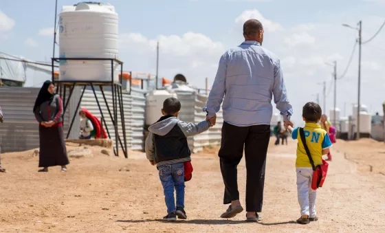 A father walks down the road of the camp he lives in with his family whilst holding his children's hands.