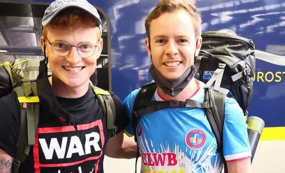 Mitch Jones wearing a War Child branded t-shirt and baseball cap smiles to the camera stood in front of a Eurostar train.