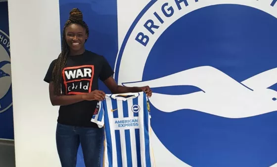 Rinsola Babjide stands in front of a wall covered in blue and white striped and the Brighton & Hove Football Club logo whilst holding her blue and white striped Brighton & Hove Football jersey. Rinsola wears a black War Child T-Shirt with War Child logo on the front.