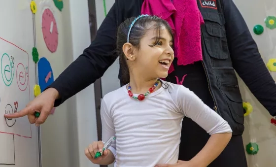 Girl smiles to her classmates as she completes a task on the whiteboard with the help of a War Child Education Facilitator in Jordan.