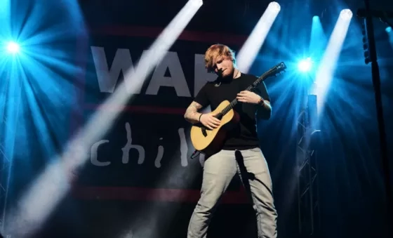 Ed Sheeran standing on a stage playing his guitar with the black, red and white War Child logo in the background. Simple white spotlights dot through the stage