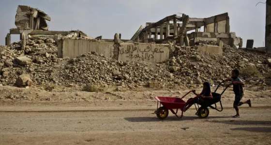 A child pushing a wheel barrow next to a destroyed building. 
