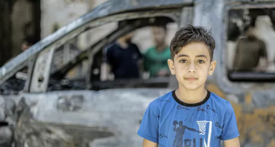 A young boy in a blue t-shirt standing in front of a car in burnt out car