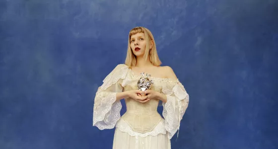 Aurora standing in front of blue wall in a white dress holding a metal heart
