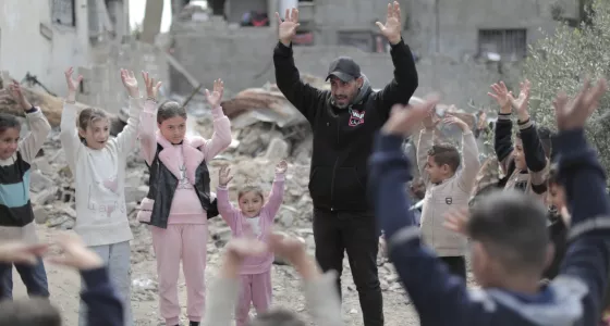 An adult man and children standing amongst rubble with hands in the air