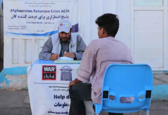 Adult man sitting behind War Child and partners branded table and an Afghan sat in a chair receiving support with his back against the camera.