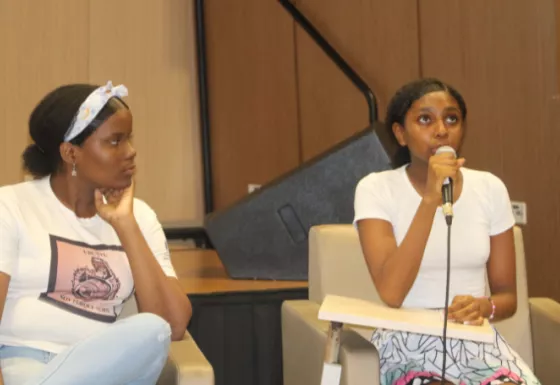 two girls sat beside each other on stage with one girl speaking whilst holding a microphone.