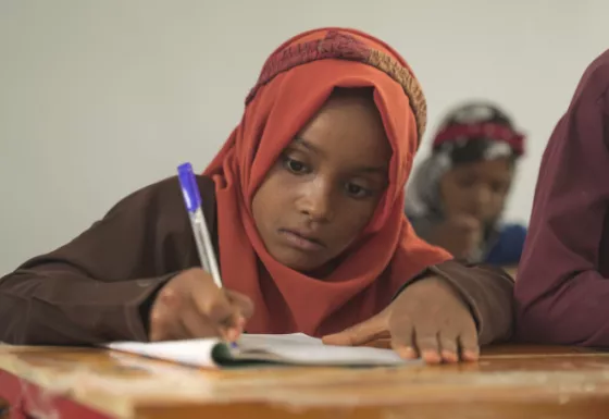 Girl wearing orange hijab sat at a table, writing with a pen.