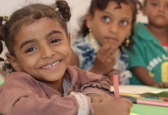 Girl sat at a desk with a pen in one hand, smiling.