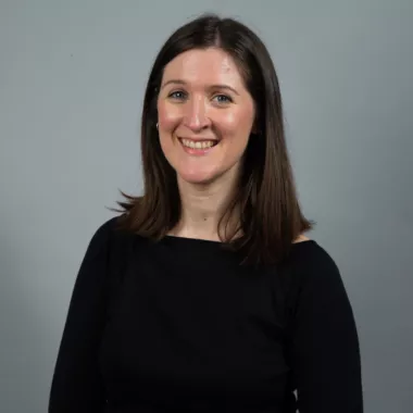 A woman standing and smiling in front of a grey background.