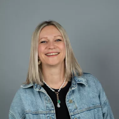 A woman standing and smiling in front of a grey background.