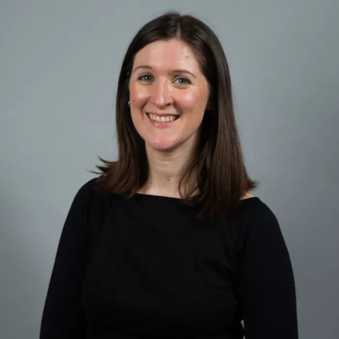 A woman standing and smiling in front of a grey background.