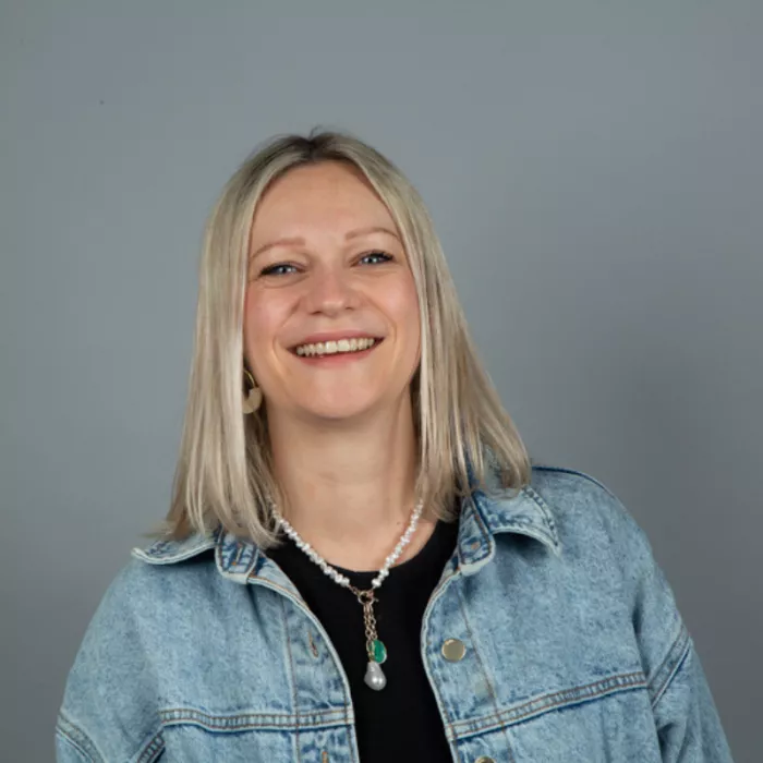 A woman standing and smiling in front of a grey background.