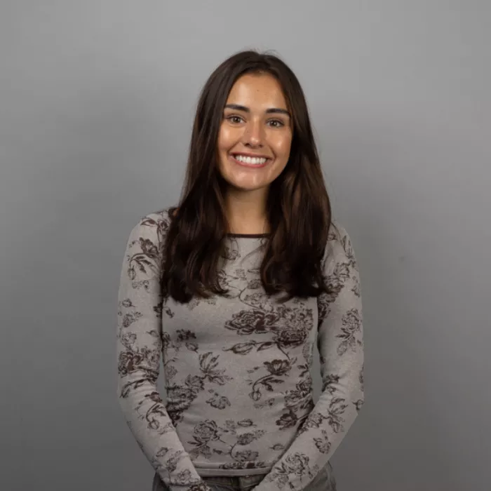 A woman standing and smiling in front of a grey background.